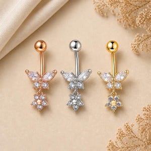 Pode incluir: Três piercings no umbigo em ouro rosa, prata e ouro. Cada anel apresenta uma barra curva com uma bola superior e um design pendurado. Os designs incluem uma forma de borboleta com cristais transparentes e um pingente de flor.