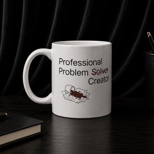 May include: A white ceramic mug featuring the black text Professional Problem Solver Creator, with the word Solver crossed out by a red line. Below the text is a graphic of a small mug spilling a brown liquid onto a stack of white papers.
