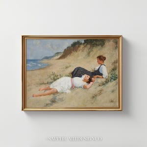May include: A framed painting depicts two figures on a sandy beach. One reclines in a white dress with a pink sash, while the other sits nearby in a dark dress. The background shows a coastline with a blue sea and a sandy cliff.