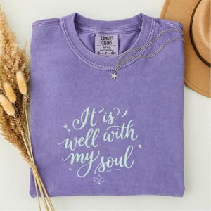 Puede incluir: Una camiseta Comfort Colors lavanda con la frase "It is well with my soul" en escritura azul claro. Un collar plateado con un colgante de estrella descansa sobre la camiseta. Un sombrero marrón y flores secas están en el fondo.