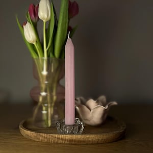 May include: A pink candle in a clear glass holder sits on a wooden tray, beside a small, pale pink flower-shaped dish. A vase of tulips with white and burgundy petals is in the background. The scene is set on a wooden surface.