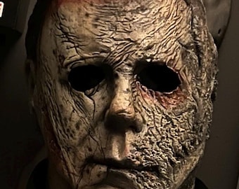 Puede incluir: Una máscara desgastada de Michael Myers con cuencas oscuras y una superficie agrietada y texturizada. La máscara tiene una apariencia realista y envejecida con áreas de decoloración y una pieza de cabello oscura y mate. Un accesorio clásico de película de terror de Halloween.