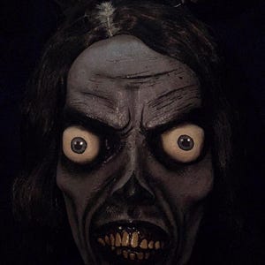 May include: A grotesque mask with a zombie-like appearance. The mask features large, yellow eyes, exposed teeth, and dark hair. The mask also has long, pointed ears, giving it a sinister look. The overall color scheme is dark and muted, creating a spooky effect.