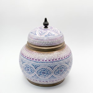 May include: A decorative ceramic jar with a lid, featuring intricate patterns in blue, purple, and white. The jar has a bronze-colored band around the middle and a black finial on top. The design is symmetrical and ornate.