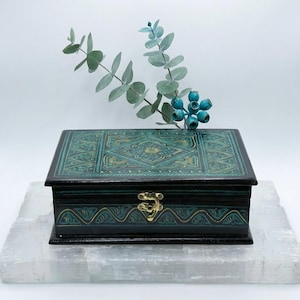 May include: A rectangular, teal and gold decorative box with a hinged lid and a gold-colored clasp. The box features intricate embossed patterns and is set on a white surface. A sprig of eucalyptus with blue berries is in the background.