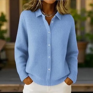 May include: A light blue collared shirt with long sleeves and a button-down front. The shirt is made of a soft, knitted material. The model is wearing white pants. The shirt has a classic design.