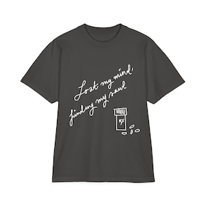 Puede incluir: Camiseta gris oscuro con texto blanco que dice "Lost my mind, finding my soul". Una ilustración blanca de un frasco de pastillas y pastillas está debajo del texto. La camiseta tiene mangas cortas y cuello redondo.