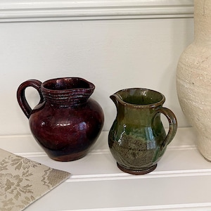 May include: A still life arrangement featuring a burgundy ceramic pitcher and a smaller green pitcher. A large, textured, off-white vase holds dried flowers. A beige floral patterned cloth is on the left.