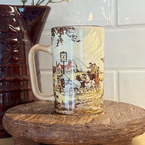May include: A cylindrical ceramic mug featuring a detailed illustration of a horse-drawn carriage traveling past a building with a sign that reads The Star. The mug has a light beige background with brown accents and a matching handle on the side.