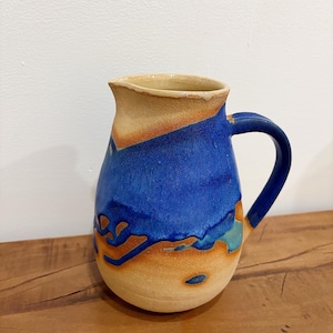 May include: A ceramic pitcher featuring a rounded body and a curved handle. The exterior displays a textured, matte beige base color with a vibrant, glossy blue glaze applied in a flowing, irregular pattern across the upper section and handle.