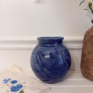 May include: A round, blue ceramic vase with a textured surface and a wide opening. The vase is next to a tan vase and a cream-colored cloth with blue floral embroidery. White flowers are in the background.