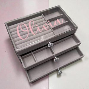 May include: A grey jewellery box with three drawers and a clear lid. The top section has ring slots. The name "Olivia" is written in pink script on the lid. Each drawer has a crystal-like knob.