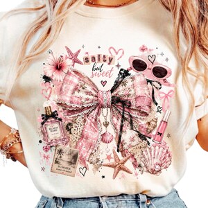 May include: A cream-colored t-shirt featuring a graphic print of a large pink plaid bow, a perfume bottle labeled Beach Babe, sunglasses, a starfish, a seashell, and a photo print labeled Summer state of mind with the text salty but sweet.