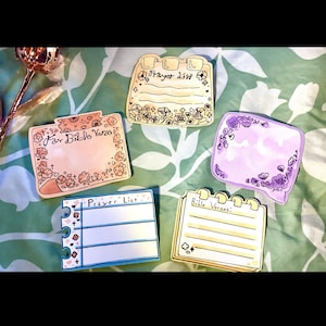 May include: Five colorful, hand-drawn notepads with floral and heart accents. The notepads have text that reads "Prayer List", "For Bible Verses", and "Bible Verses". The notepads are on a green and white floral patterned background.