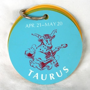 May include: A circular card stack bound by a silver metal ring. The top card is light blue with a red illustration of a bull, the text "APR. 21-MAY 20" at the top, and the word "TAURUS" printed in white capital letters at the bottom.