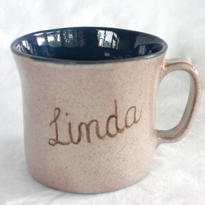 May include: A ceramic mug with a speckled beige exterior and a dark blue interior. The name Linda is written in brown cursive script on the front of the mug. It features a single handle on the right side and a smooth, rounded rim.