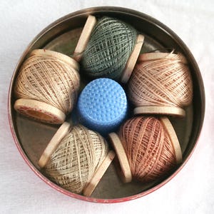 May include: A vintage sewing kit featuring a red tin container holding several spools of thread in various neutral colors, a blue thimble, and wooden spool holders. The spools are filled with thread, and the thimble is in the center.