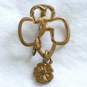 May include: A gold-toned metal pin featuring a stylized figure of a person in a pointed hat centered within a clover-like frame. A small, separate five-petaled flower charm hangs from the bottom of the main frame by a small metal jump ring.