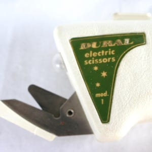 May include: Vintage DURAL electric scissors, model 1, with a white body and silver blades. The green label features gold lettering and three stars. The scissors are designed for cutting fabric or paper.