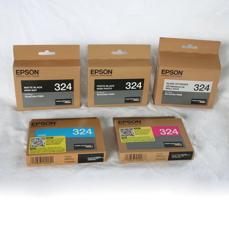 May include: Several Epson ink cartridges in cardboard boxes. The boxes are labeled with the Epson logo and product information, including "Matte Black," "Photo Black," "Gloss Optimizer," and "Cyan." Each box displays the number "324."