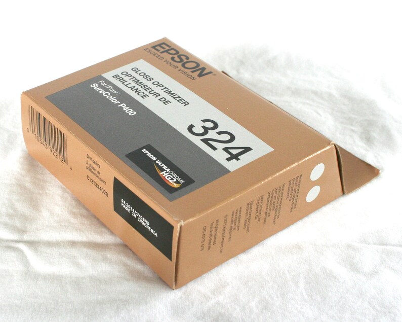 K&ouml;nnte beinhalten: Ein brauner Karton mit dem Text "EPSON EXCEED YOUR VISION" und "GLOSS OPTIMIZER OPTIMISEUR DE BRILLANCE" auf der Vorderseite. Der Karton hat auch den Text "For Pour: SureColor P400" und "324" auf der Vorderseite. Der Karton hat ein schwarzes Etikett mit dem Text "EPSON ULTRACHROME HG2" darauf.