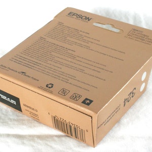 K&ouml;nnte beinhalten: Ein brauner Karton mit einem wei&szlig;en Etikett, auf dem "EPSON" und "Better Products for a Better Future" steht. Die Schachtel enth&auml;lt zwei Tintenpatronen f&uuml;r einen Drucker. Die Schachtel hat auch einen Barcode und den Text "2 Cartridges" und "14ml/cartridge".
