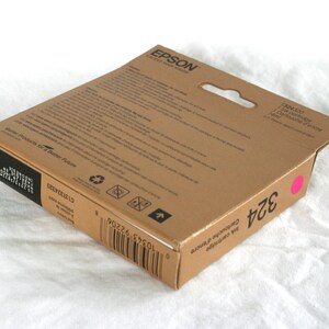 May include: A brown Epson ink cartridge box with the number 324 in black and a pink dot. The box has the text "Ink Cartridge" and "Better Products for a Better Future".