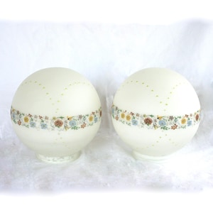 May include: Two off-white glass globe lampshades. Each shade features a floral band with blue, yellow, and brown flowers. Small green dots are arranged in rows across the surface. The lampshades have a rounded base.