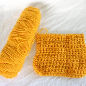 May include: A close-up shot of a skein of bright yellow yarn next to a small, knitted swatch. The yarn is tightly wound, and the knitted piece shows a textured pattern. The background is a plain white fabric.
