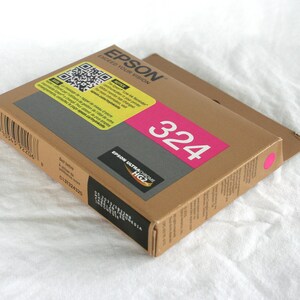 May include: A brown cardboard box of Epson 324 ink cartridges. The box has a pink label with the number 324 in white. The Epson logo and "Exceed Your Vision" are printed on the top. The box is on a white surface.
