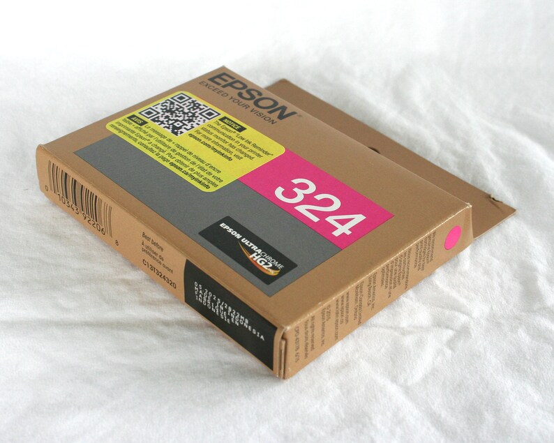 May include: A brown cardboard box of Epson 324 ink cartridges. The box has a pink label with the number 324 in white. The Epson logo and "Exceed Your Vision" are printed on the top. The box is on a white surface.