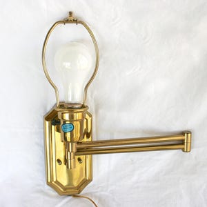 May include: A vintage brass wall-mounted lamp with an adjustable arm. The lamp features a large, frosted light bulb and a decorative, geometric base. The arm extends outward, allowing for adjustable positioning. A small blue sticker is visible on the base.