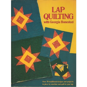 May include: Book cover with the title "LAP QUILTING with Georgia Bonesteel". The cover features a yellow background with three quilt blocks in the foreground. The quilt blocks are made of green, yellow, and red fabric and feature a star design.