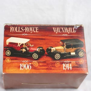 May include: A vintage toy car box featuring two classic car models: a red Rolls-Royce from 1906 and a gold Vauxhall from 1914. The box has a brown wooden-like background with the car names in bold text.