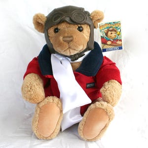 May include: A plush teddy bear wearing a brown aviator helmet and goggles, a red jacket with a navy blue collar, and a white scarf. The bear has a tag that reads "Lands' End" with an illustration of a bear in a plane.
