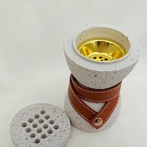 Può includere: Un bruciatore di incenso bianco maculato con un cinturino in pelle marrone e un interno color oro. Il bruciatore ha un coperchio rimovibile con più fori. Il design è moderno e minimalista, adatto all'arredamento della casa.