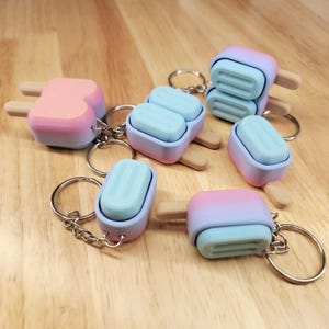 May include: A collection of popsicle-shaped keychains featuring ombre color gradients in shades of pink, blue, and purple. Each keychain includes a metal ring and a small tan stick, with some designs showing single or double popsicle segments.
