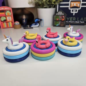 May include: A collection of six small, circular, multi-colored plastic pool float toys. Each toy features a miniature animal figure, including unicorns, pink flamingos, and yellow ducks, resting on top of stacked rings in shades of blue, yellow, and pink.