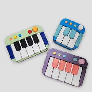 May include: Three colorful toy pianos. One is green with black and white keys, another is blue with white and light blue keys, and the third is purple with white and pink keys. Each has a different color scheme and button layout.
