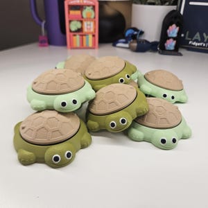 May include: A collection of small, 3D-printed turtle-shaped fidget toys. Each turtle features a light green or olive green body with a textured, tan-colored shell and two prominent white eyes with black pupils, arranged in a cluster on a surface.