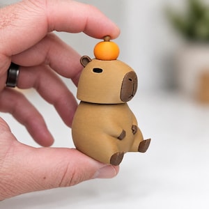 May include: A small, tan, capybara-shaped figurine with a dark brown face and feet. The capybara is sitting in an upright position and has a small, round, bright orange sphere balanced on the top of its head. The surface has a matte texture.