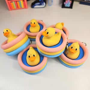 May include: A collection of five yellow rubber duck keychains, each sitting inside a miniature inflatable pool. The pools feature a blue base with a pink and yellow ringed rim. Each duck has a bright orange beak and black eyes, with a metal keychain ring.