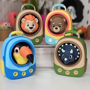 May include: Four small, colorful, backpack-shaped containers, each featuring a different animal head peeking out from a circular opening. The animals include a lion, a bear, a toucan, and a triceratops. Each container has a handle and three circular accents at the base.