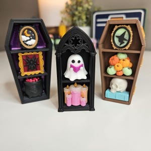 May include: Three miniature display shelves featuring two coffin-shaped units and one arched gothic-style unit. The shelves contain small decorative items including a ghost reading a book, a skull, pumpkins, candles, a spider, and a cauldron.