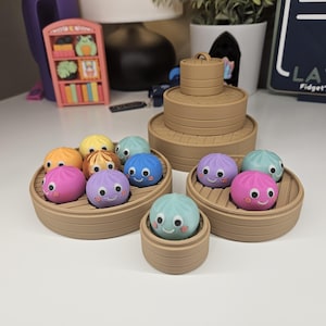 May include: A collection of small, round, colorful silicone dumpling toys with googly eyes and smiling faces, each resting inside a tan, circular, tiered plastic steamer basket container. The set includes various sizes of stacked and individual steamers.