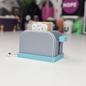 May include: A gray plastic toaster-shaped keychain with a light blue base and a lever on the side. A tan pastry with white frosting and colorful sprinkles is inserted into the top slot. A white ball chain is attached to the side of the toaster.