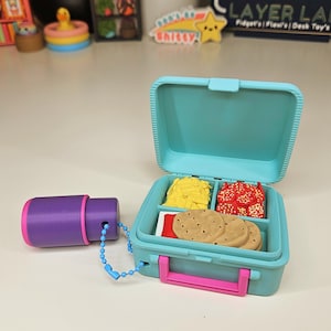 May include: A light blue plastic lunch box with a pink latch, containing miniature food items including yellow fries, red pepperoni slices, and three tan cookies. A purple cylindrical container is attached by a blue ball chain to the lunch box.