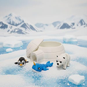 May include: A miniature arctic scene featuring a white igloo with an open lid, a black and white penguin, a blue narwhal with a yellow horn, and a white polar bear. The scene is set on a snowy, icy surface.