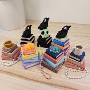 May include: A collection of miniature book-themed charms. Each charm features a stack of colorful books topped with various designs, including a black crow, a skull, a sleeping cat, a dragon, and a coffee cup. Each charm has a beaded chain.