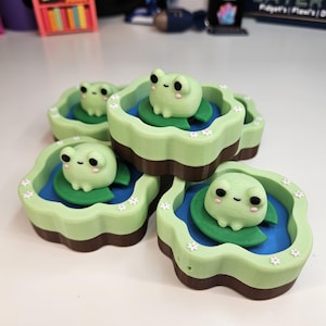 May include: A collection of four small, 3D-printed containers, each featuring a light green, scalloped-edge rim decorated with tiny white flowers. Inside each container is a blue pond base with a green lily pad and a small, light green frog.